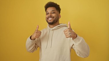Gülümseyen Afrikalı Amerikalı adam sarı bir arka plana karşılık günlük giysiler içinde başparmağını kaldırıyor..