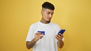 Genç, yakışıklı Latin bir adam, dövmeli ve üzgün, rastgele kredi kartını ve akıllı telefonunu izole edilmiş sarı duvara karşı kullanıyor..