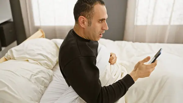 A surprised hispanic man in a bedroom holding a phone with a shocked expression on his face, depicting an indoor morning scenario.