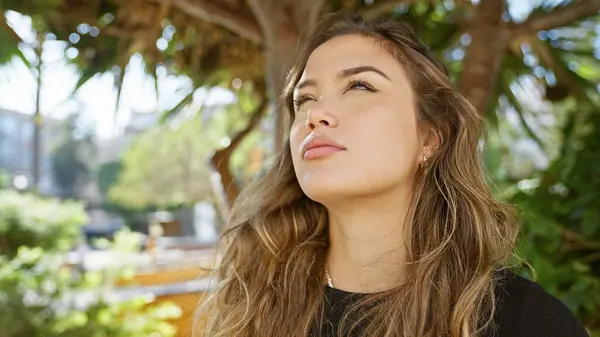 Young, relaxed hispanic woman with a serious expression, looking up at the sky in a sunlit park, radiating natural beauty in a casual lifestyle portrait,