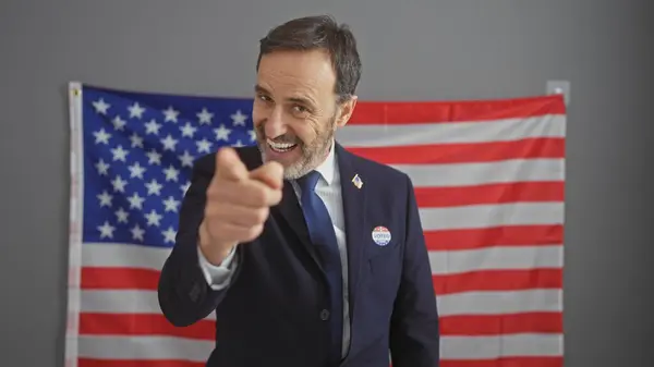 Smiling bearded man in suit pointing with american flag behind, symbolizing leadership and patriotism