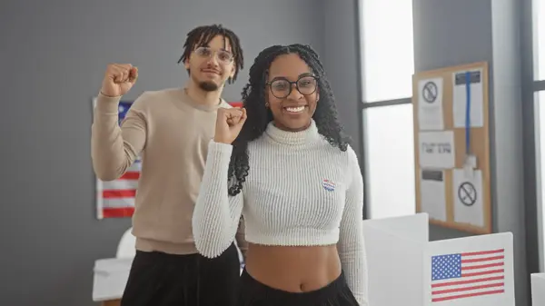 stock image A cheerful man and woman with 'i voted' stickers, raising their fists in unity at an american polling station, symbolizing democratic participation.