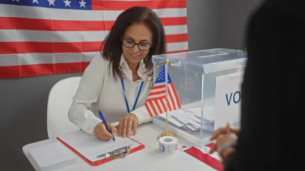 stock image A woman volunteering at a united states electoral college writes notes under an american flag indoors.