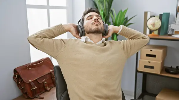 stock image A relaxed young hispanic man with a beard enjoys music with headphones in a modern office setting.