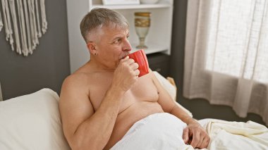 Tişörtsüz, orta yaşlı bir adam, rahat yatak odasında kırmızı bir fincan kahveden zevk alır. Rahatlamış bir sabahı örnek olarak gösterir..