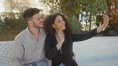 A smiling woman and man take a selfie together in an outdoor park setting, portraying a happy couple moment. clipart