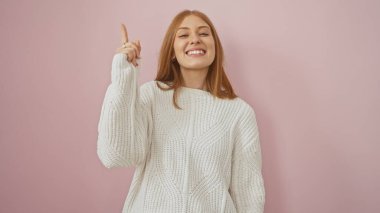 Smiling young woman with redhead in a white sweater posing against a pink background, gesturing with hand. clipart