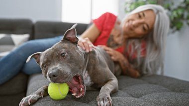 Orta yaşlı, kır saçlı, köpeği sırıtan, kendine güvenen, evinde oynayan bir kadın.