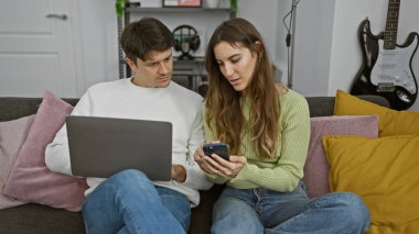 Modern bir oturma odasında renkli yastıklarla kanepede oturan bir kadın ve erkek telefon ve dizüstü bilgisayar kullanıyor..