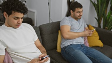 Modern bir oturma odasında gri bir koltukta oturan iki adam akıllı telefonları karıştırıyor..