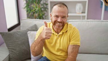 Kendinden emin, gülümseyen, orta yaşlı, beyaz bir adam başparmağını kaldırıyor, oturma odasındaki kanepesinde rahatça oturuyor, evinde neşe ve rahatlık saçıyor.