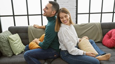 Gülümseyen bir çift, rahat bir oturma odasında sırt sırta oturur, rahat bir yuva ortamında sevgi ve arkadaşlık yayarlar..