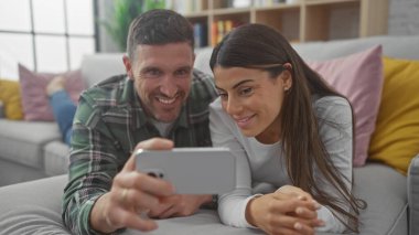 Gülümseyen İspanyol bir çift evdeki rahat oturma odalarında bir kanepede selfie çekiyorlar..