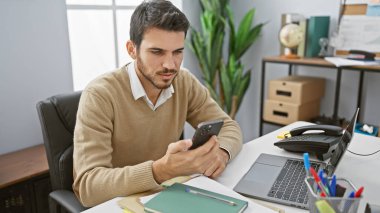 Ofiste çalışan, odaklanmış, sakallı genç bir İspanyol iş için akıllı telefonunu ve laptopunu kullanıyor..