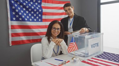 Two smiling women conducting electoral process in an american college with a prominent flag and voting box in an indoor setting. clipart