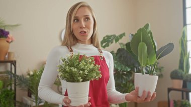 Kırmızı önlüklü şaşırmış sarışın bir kadın iyi aydınlatılmış bir çiçekçinin içinde saksı saksısı tutuyor..