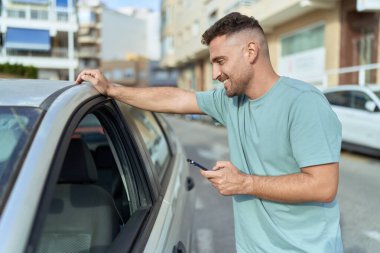 Young hispanic man using smartphone leaning on car at street