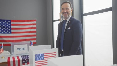 Amerikan bayraklarıyla bir Amerikan seçim merkezinde gülümseyen sakallı olgun bir adam..