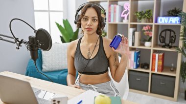 Young woman with headphones in a radio studio holding a blue pill bottle, showcasing a health segment. clipart