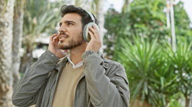 Sakallı, genç, yakışıklı bir İspanyol, açık havada, serin bir parkta, yemyeşil bir yerde kulaklıkla müzik dinlemekten hoşlanıyor..