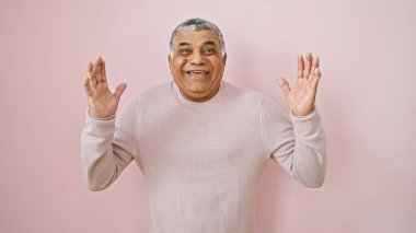 Beyaz saçlı, neşeli orta yaşlı bir adam pembe bir duvarın önünde el kaldırarak el kol hareketi yapıyor..