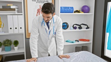 Beyaz laboratuvar önlüklü İspanyol bir adam klinik rehabilitasyon odasında duruyor. Üzerinde tıbbi posterler ve ekipmanlar var..