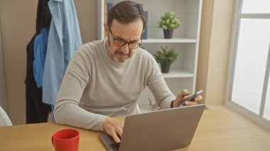 Sakallı, orta yaşlı bir adam akıllı telefon ve dizüstü bilgisayar kullanıyor..
