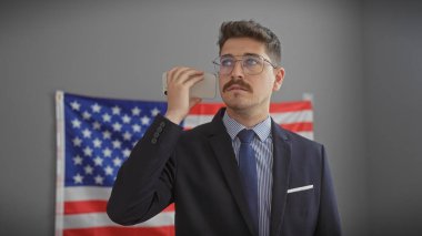 Yakışıklı İspanyol takım elbiseli adam ofisinde Amerikan bayrağıyla akıllı telefon kullanıyor.