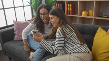 İki kadın, anne ve kız, rahat bir oturma odasında akıllı telefona bakarken neşeyle gülüyorlar..