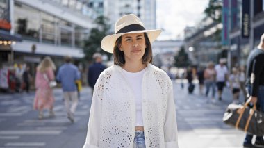 Güzel, genç, İspanyol bir kadın ciddi bir ifadeyle Stockholm sokaklarında yaz şapkası takmış kameraya bakıyor.