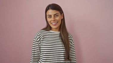 Çizgili tişörtlü gülümseyen genç bir İspanyol kadın pembe izole bir arka plana karşı duruyor. Güzellik ve pozitifliği somutlaştırıyor..