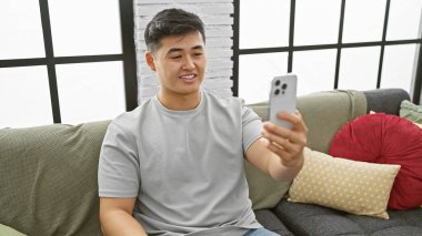 Genç Asyalı bir adam akıllı telefonuyla şık bir oturma odasında selfie çekiyor.