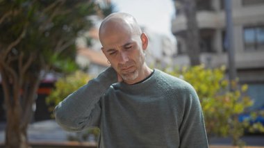 Sakallı dalgın kel bir adam yeşil bir şehir parkında dikilirken rahatsız bir şekilde boynuna dokunuyor..