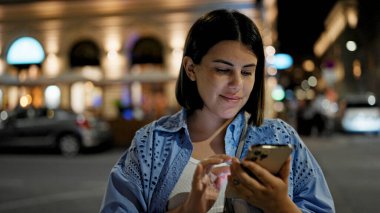 Genç ve güzel İspanyol kadın gülümseyerek mutlu bir şekilde sokaklarda akıllı telefon kullanıyor.