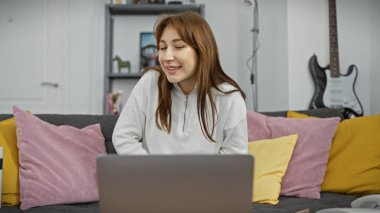 Oturma odasında renkli yastıkları olan bir kanepede dizüstü bilgisayar kullanan gülümseyen genç bir kadın..