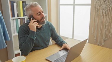 Laptop kullanan ve modern bir oturma odasında telefonla konuşan düşünceli sakallı bir adam..