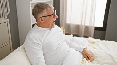 Beyaz gömlekli, gözlüklü, orta yaşlı, gülen bir adam bir apartman dairesinde oturuyor.
