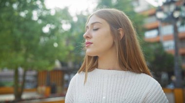 Sakin, gözleri kapalı genç bir kadın sakin bir şehir parkında, doğanın ortasında duruyor..