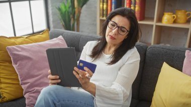 Elinde kredi kartı ve tablet olan olgun İspanyol bir kadın. Kapalı alandaki koltukta oturuyor..