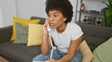 Düşünceli bir Afrikalı kadın modern oturma odasında telefonla konuşuyor, sakin bir ortam yayıyor..