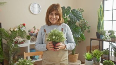 Gülümseyen kadın çiçekçide çiçek tutuyor. Etrafı yeşillik ve çiçeklerle çevrili. Kapalı alanlarda doğal hobileri var..