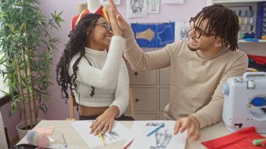 Erkek ve kadın terziler, eskizler ve dikiş ekipmanlarıyla çevrili iyi donanımlı bir atölyede başarılı bir tasarımı kutluyorlar..