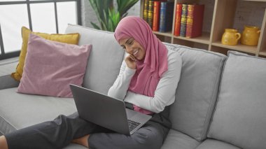 Canlı yastıklar ve kitaplarla dolu rahat bir oturma odasında dizüstü bilgisayar kullanan tesettürlü bir kadın..