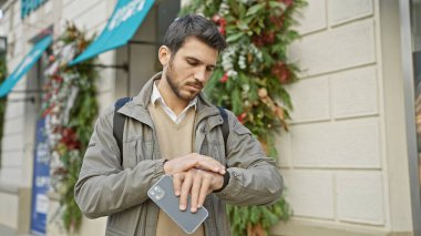 Yakışıklı İspanyol bir adam şehir caddesinde akıllı telefon tutarken bilek saatini kontrol ediyor..