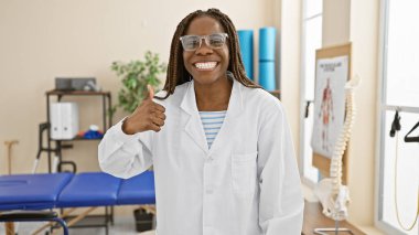 Laboratuvar önlüklü, örgülü Afrikalı Amerikalı bir kadın klinikte baş parmağını kaldırıyor..