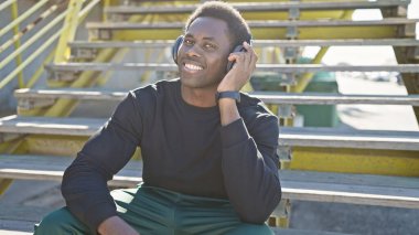 Kulaklıklı gülümseyen genç adam günlük kıyafetlerle şehir basamaklarında rahatça oturur ve güneşli havanın tadını çıkarır..