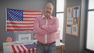 Kollarını kavuşturmuş kendinden emin kıdemli bir adam bayrak ve oy sandığıyla bir Amerikan seçim merkezinde duruyor..