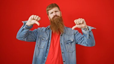 Cool, bearded redhead guy giving thumbs down  a negative sign of failure in casual lifestyle fashion, standing over an isolated red wall  clipart