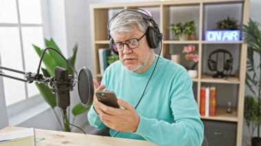Radyo stüdyosunda kulaklık takan gri saçlı olgun bir adam akıllı telefona bakıyor.