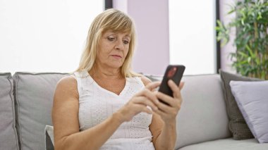 Ciddi orta yaşlı sarışın kadın teknolojiyle boğuşuyor, koltukta otururken üzgün bir ifade takınıyor, akıllı telefona mesaj yazıyor, şık oturma odasındaki sorunla boğuşuyor.